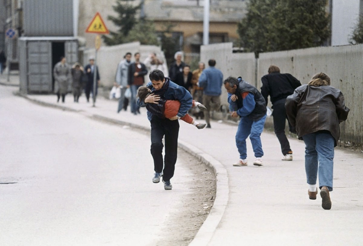 A Bosnian man cradles his child as they and others run past one of the worst spots for snipers that pedestrians have to pass in Sarajevo, on April 11, 1993.