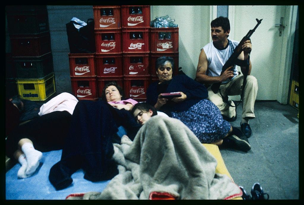 People lie on beds and sit on the floor while a man holds a rifle in a shelter during bombings in Osijek.