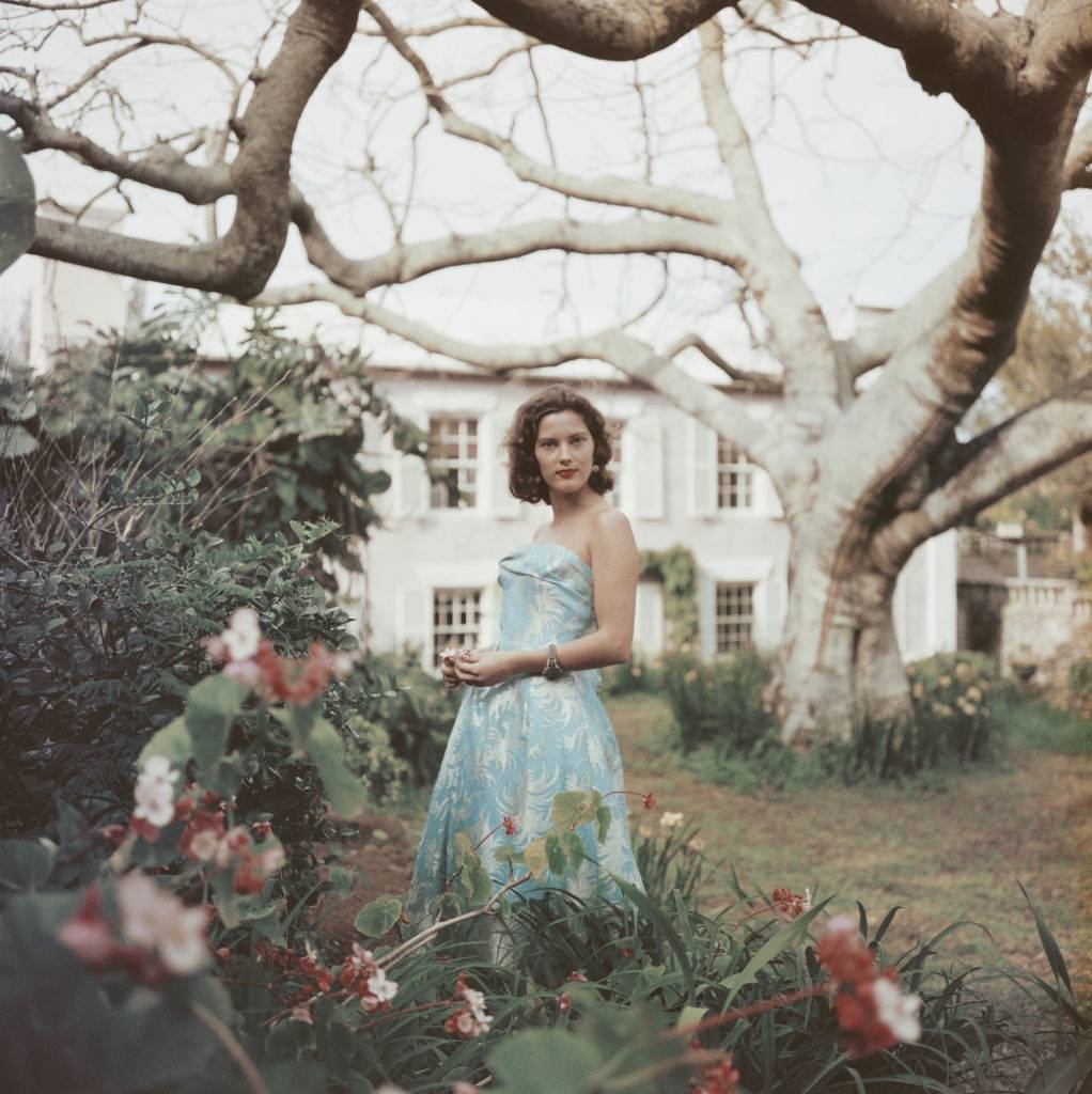 An unidentified woman stands in a garden wearing a strapless blue dress, Bermuda, 1957.