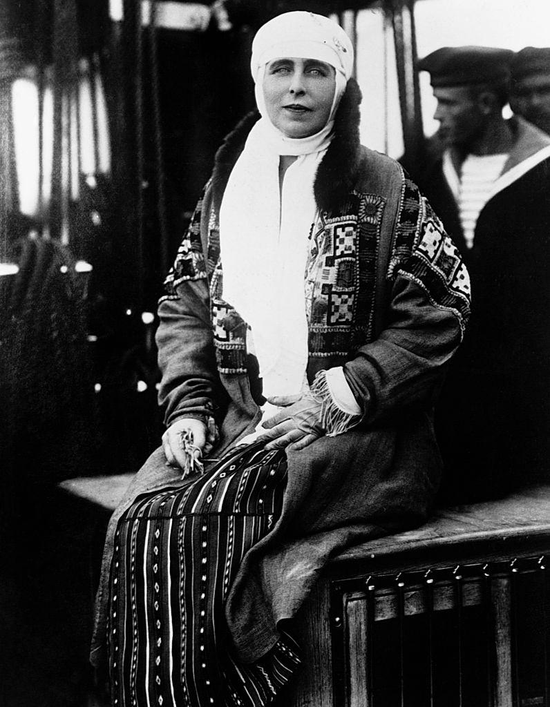 Queen Marie of Romania sits on a case while a sailor stands behind her.