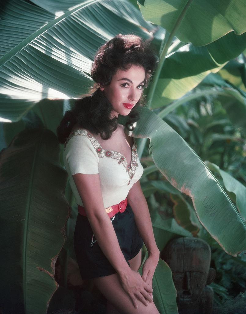 Rita Moreno in white top and black shorts posing near a tree, 1954.