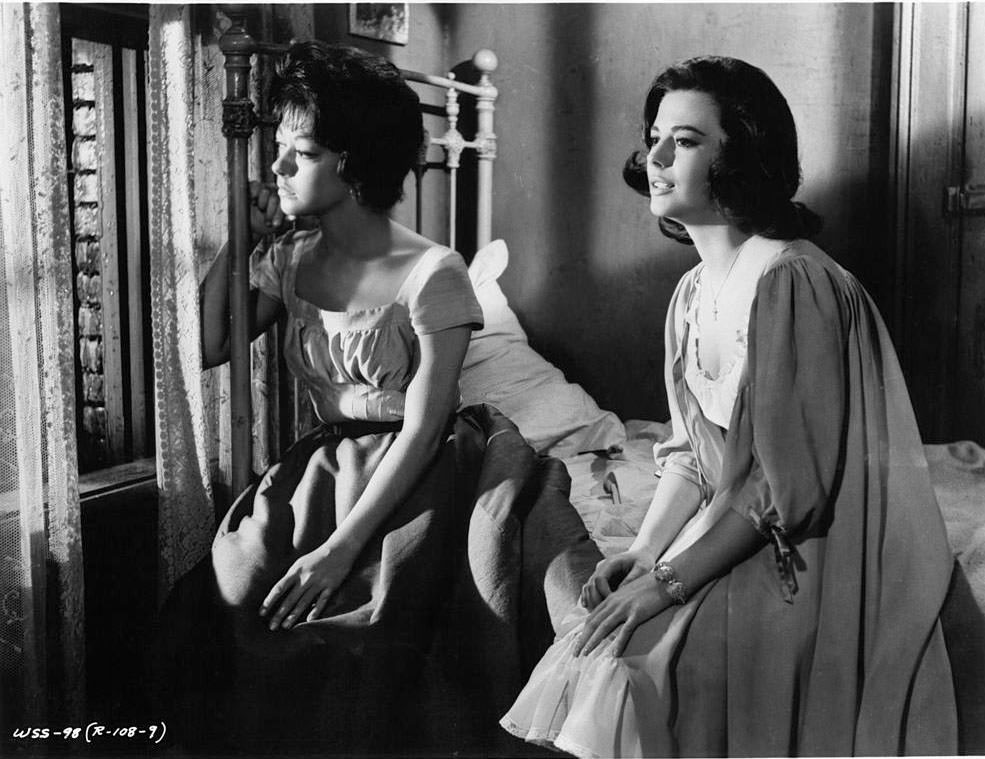 Rita Moreno and Natalie Wood sitting on a bed looking out of a window together in a scene from the film 'West Side Story', 1961.