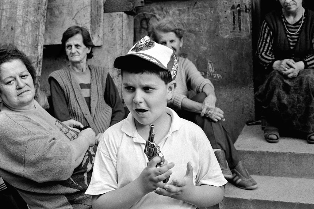 Mothers sit talking outside an apartment block during a break in shelling as a child holds a toy gun nearby.