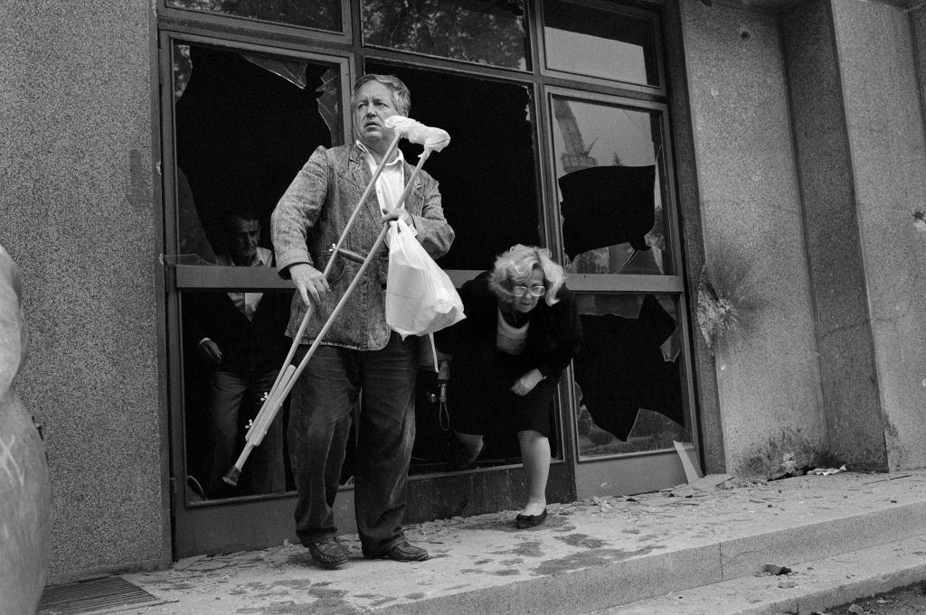 A man holds a crutch as he waits for his wife to climb through a broken shop window on Sniper Alley, July 1992.