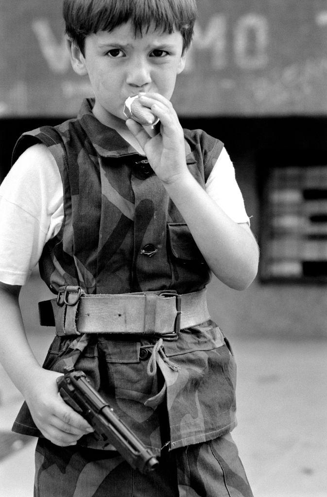 A small boy dressed in camouflage uniform holds his fathers pistol while eating an ice cream, 1992.