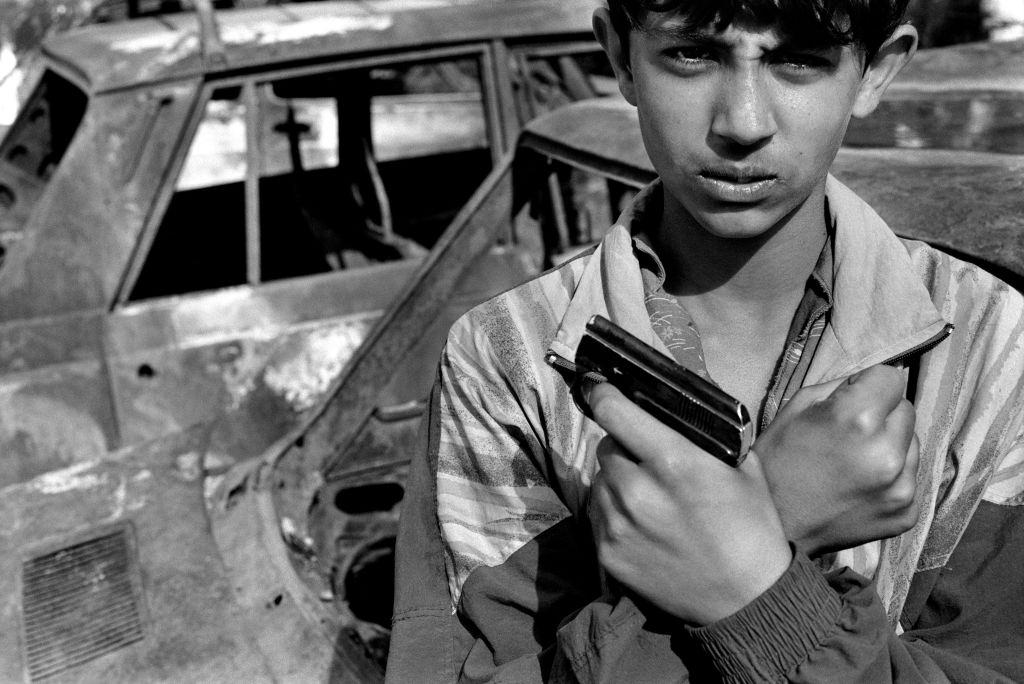 A portrait of a boy holding a small automatic pistol beside destroyed cars in a frontline suburb of the city, 1992.