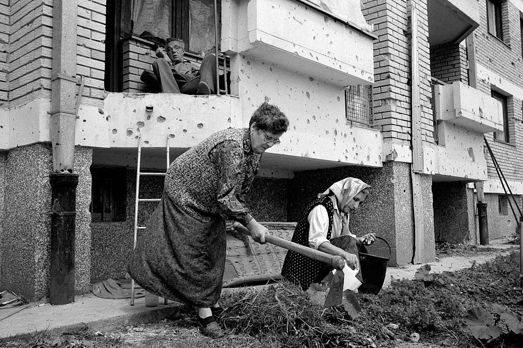 Muslim women plant vegetables in a small patch of earth outside their apartment as a man watches, and smokes a cigarette, 1994.