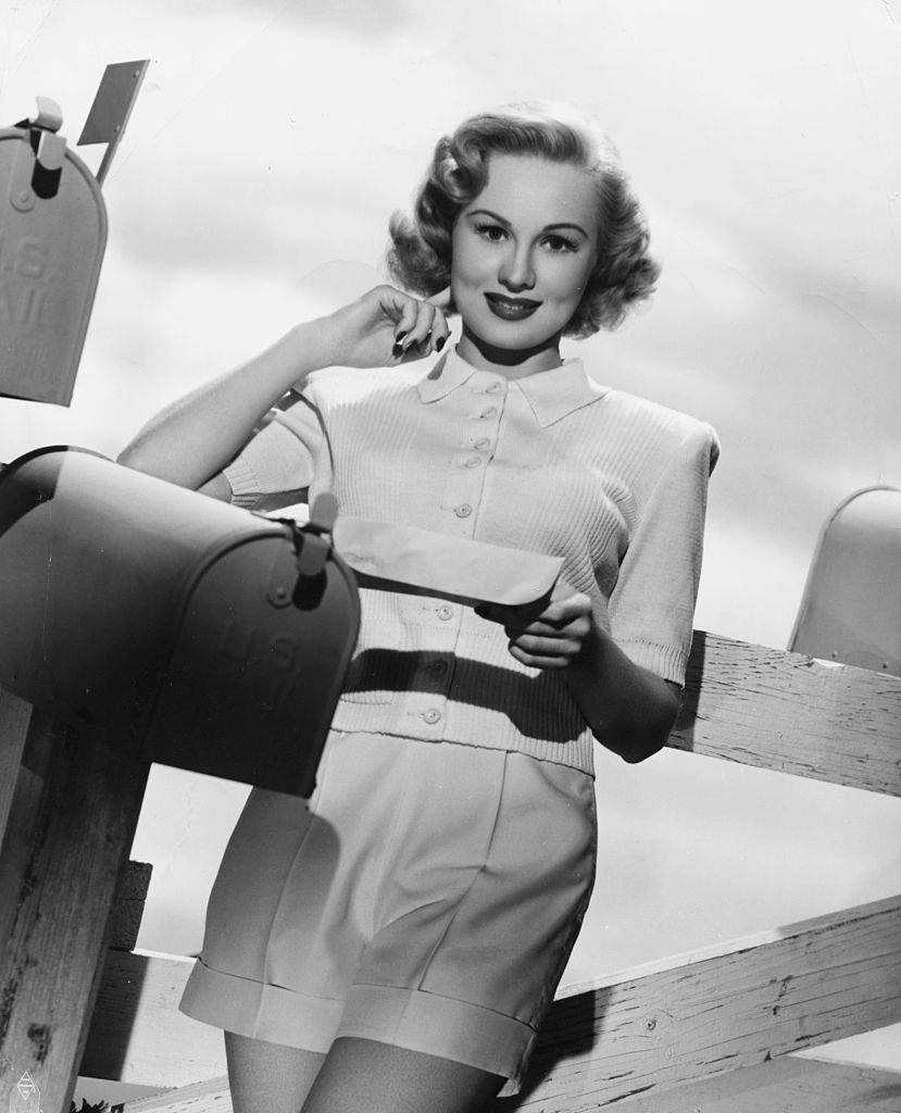 Virginia Mayo leaning on a fence and reading mail from a mailbox, 1945.