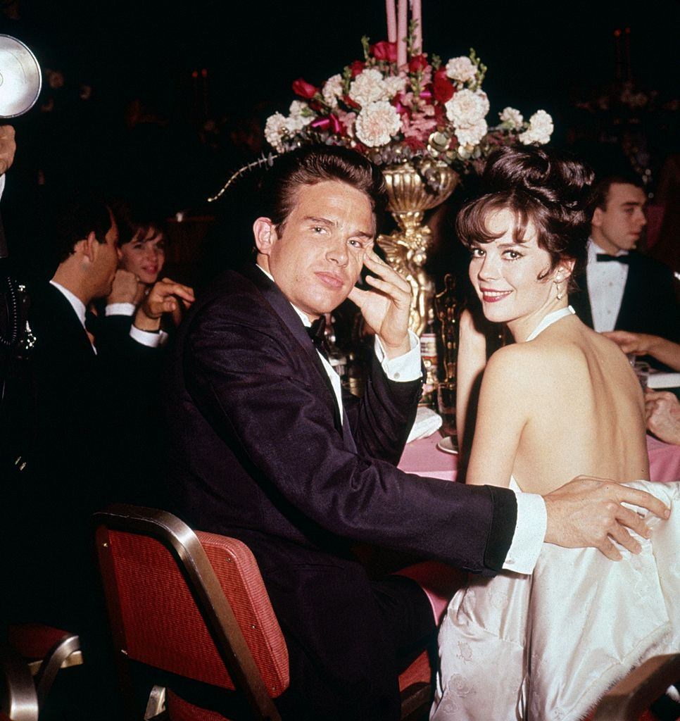 Warren Beatty and Natalie Wood look over their shoulders while seated together at a formal event, 1961.