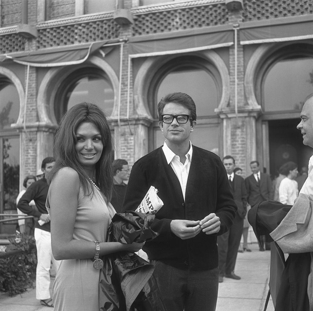Warren Beatty, wearing a cardigan and glasses, portrayed outside the Excelsior Hotel while standing next to a woman holding a newspaper, 1965.