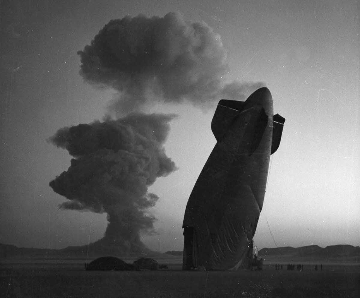 At a Nevada nuclear test site test Site, on August 7, 1957, the tail of a U.S. Navy Blimp is photographed with the cloud of a nuclear blast in the background.