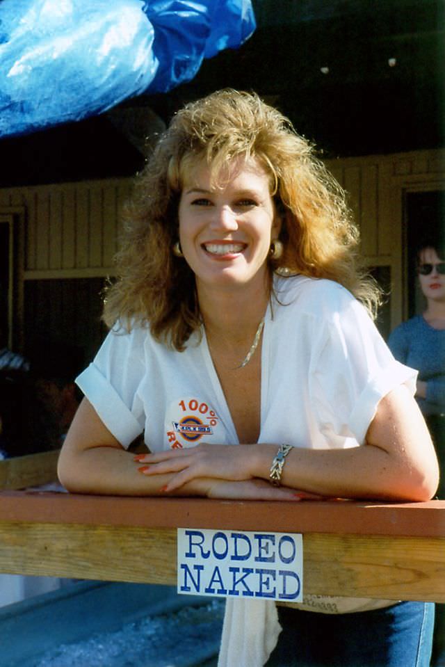 A woman working a drink booth poses by a sign reading "Rodeo Naked" during a festival, possibly the Chisholm Trail Roundup, Fort Worth Stockyards, June 1994