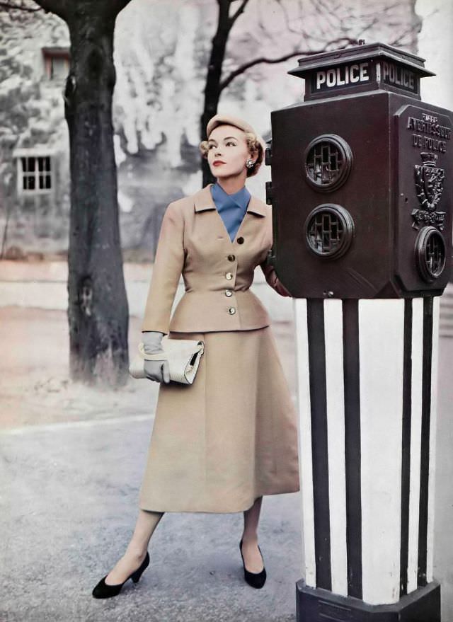 Joan Olson in classic beige suit, the skirt is bell-shaped, jacket has nipped waist and low neck that shows a bright blue scarf by Jean Patou, 1954
