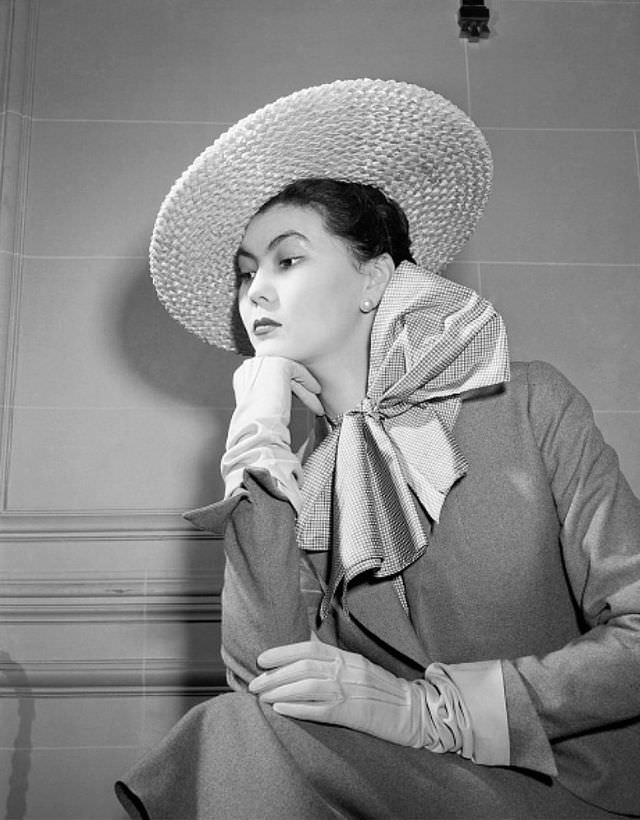 Alla in a simple gray suit with a checked blouse featuring a huge bow at the throat, natural straw sailor hat tilts over one eye, 1949