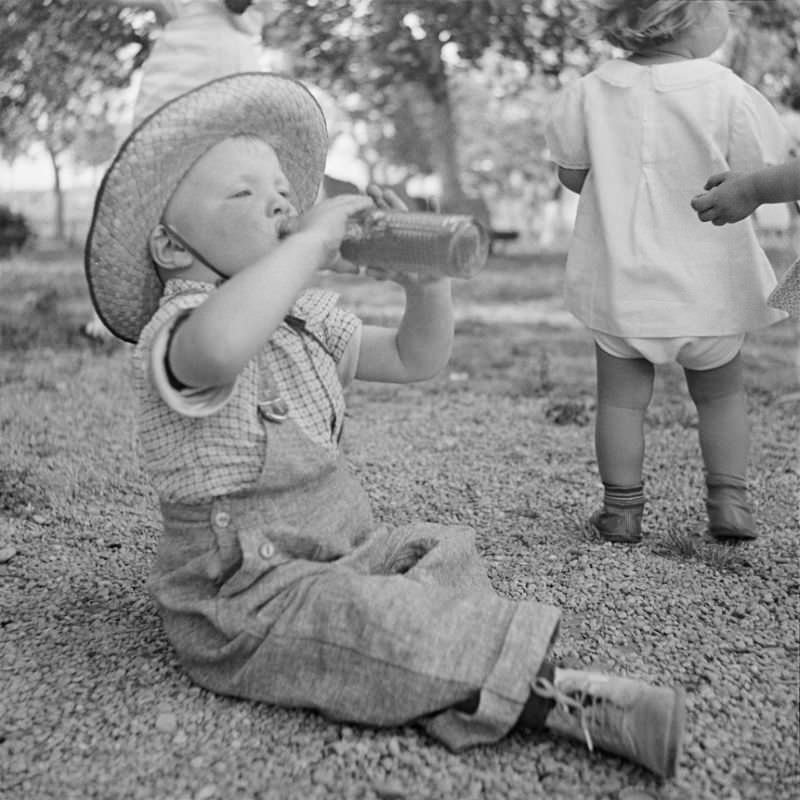 Little boy sitting on the ground, wearing a hat and drinking from a bottle