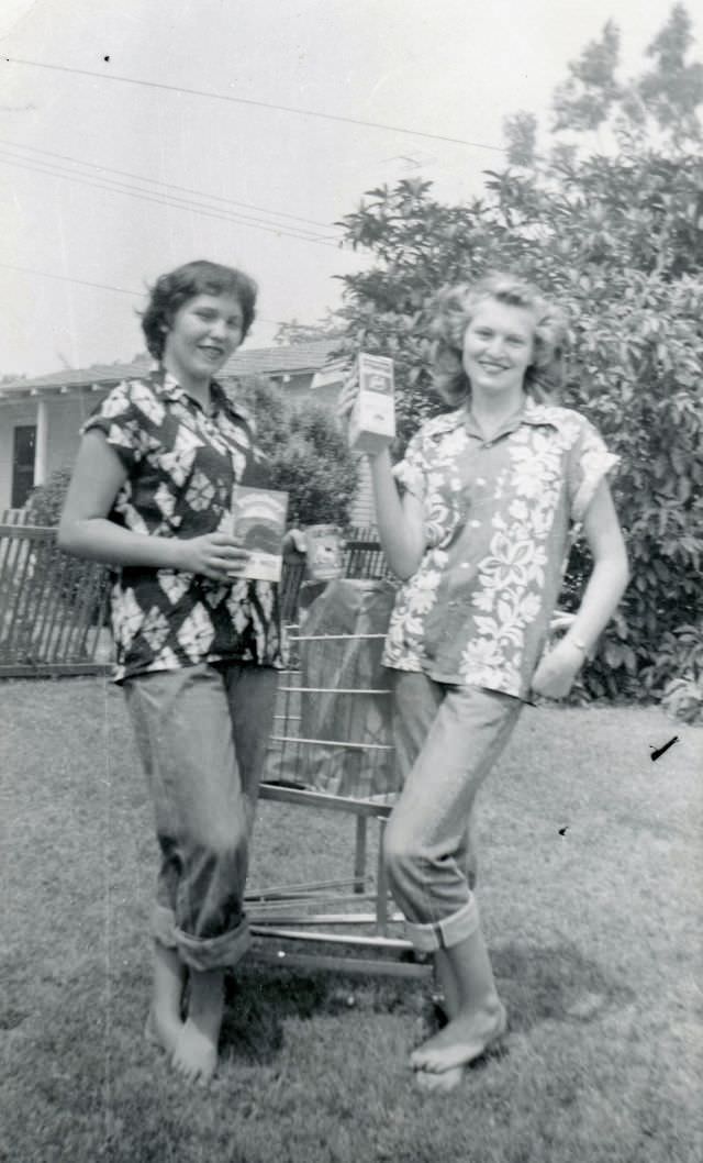 Two girls wear Hawaiian shirts, rolled jeans and are barefoot, and a shopping cart with a bag full of more products are behind them