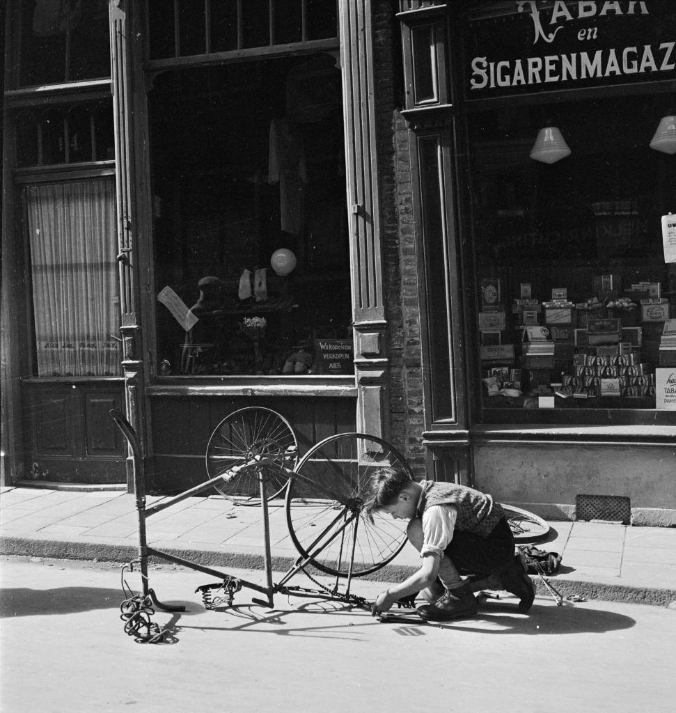 A boy repairing an old-fashioned bicycle on a road in Amsterdam, 1946.