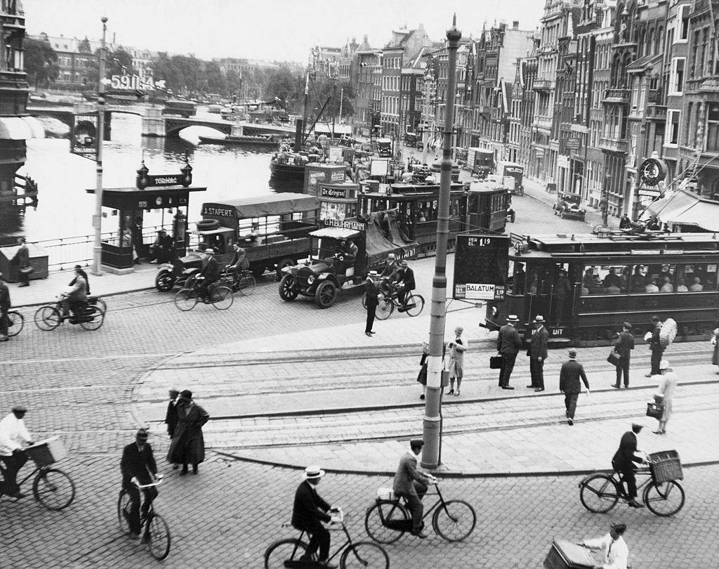 Traffic scene on one of the principal streets of the Dutch capital. It includes bicycles, automobiles, trucks, trolleys, buses, pedestrians, and freight boats on the canal, 1950s.