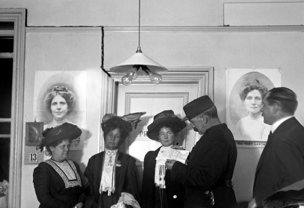 Suffragettes Flora Drummond, Emmeline Pankhurst and Christabel Pankhurst, listening to Inspector Jarvis reading an arrest warrant.
