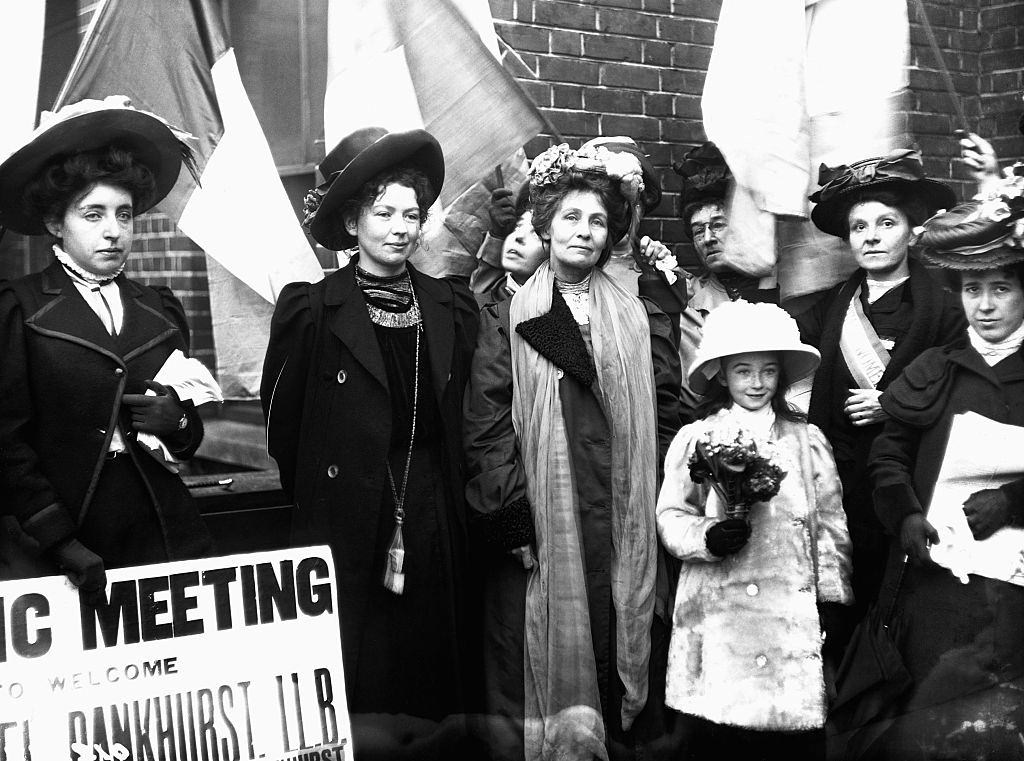 Emmeline Pankhurst and one of her daughters are welcomed to a meeting of fellow suffragettes with banners and flowers.