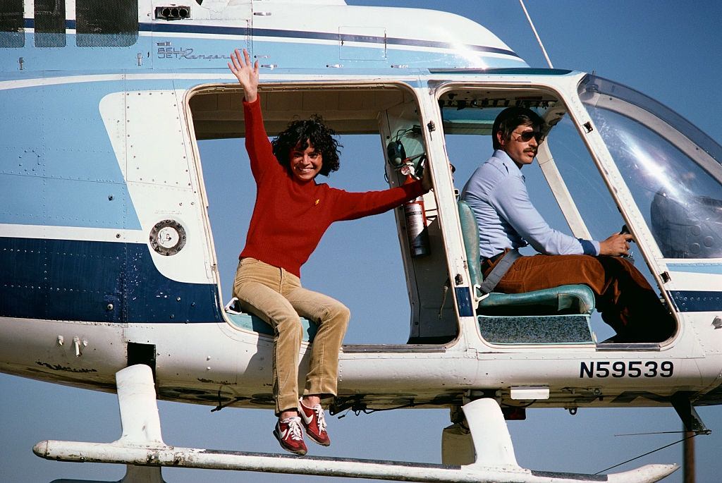 A stunt performer, playing Lynda Carter in Wonder Woman, waves from a helicopter before jumping.