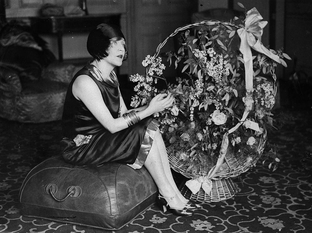 Mae Murray seated on a pouffe with a large basket of flowers, 1925.