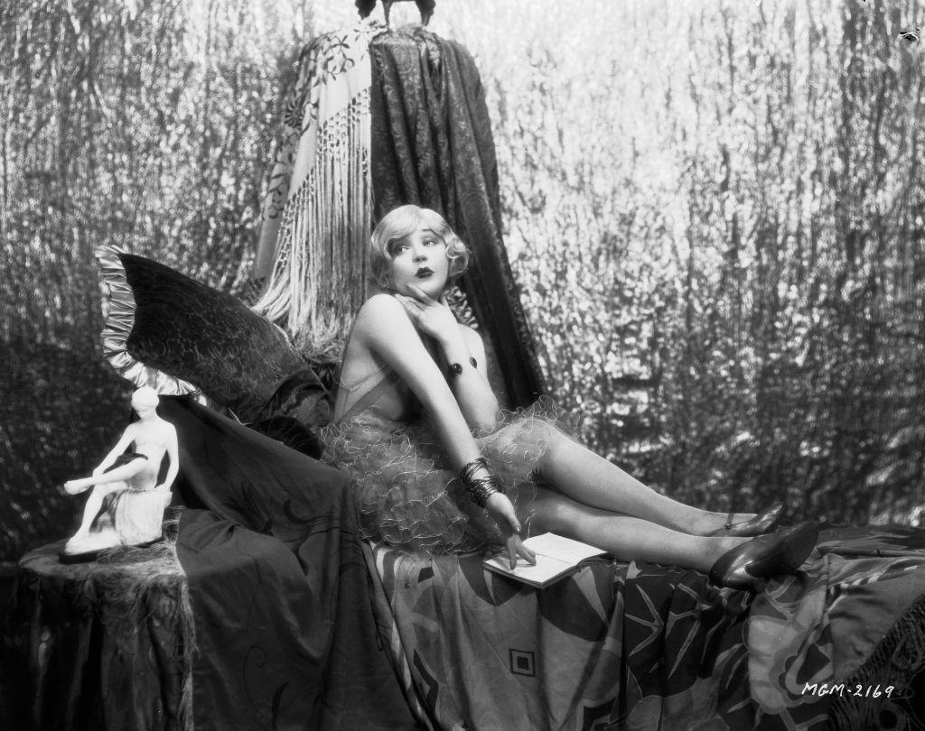 Mae Murray poses with a book against a backdrop of draperies, 1926.