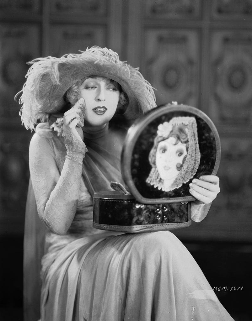 Mae Murray making herself up in a mirror in the lid of her make-up box, 1926.
