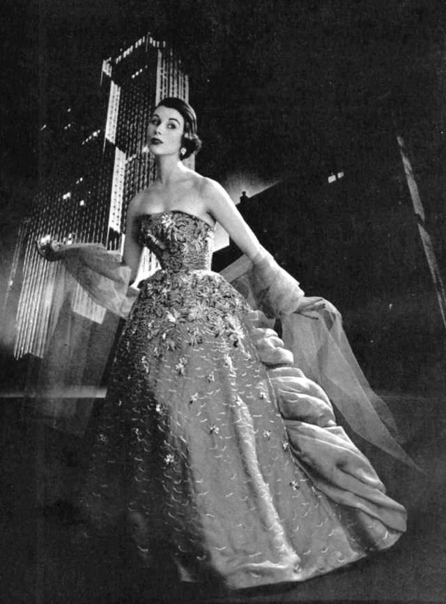 Myrtle Crawford is wearing gala ball gown of dark gray organdy embroidered with frosted multi-colored sequins, worn with light gray tulle stole by Jacques Fath, 1953