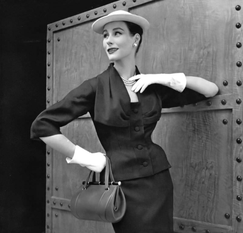 Myrtle Crawford in navy blue wool afternoon suit, jacket has a large cowl neck held by a grosgrain buttoned placket, by Lanvin-Castillo, handbag by Violette Cornille, 1954