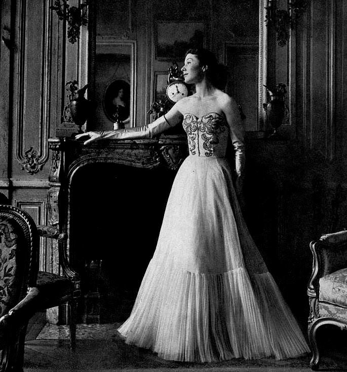 Bettina Graziani is wearing an evening dress of white tulle with finely pleated flounce, the strapless bodice is embroidered in gold