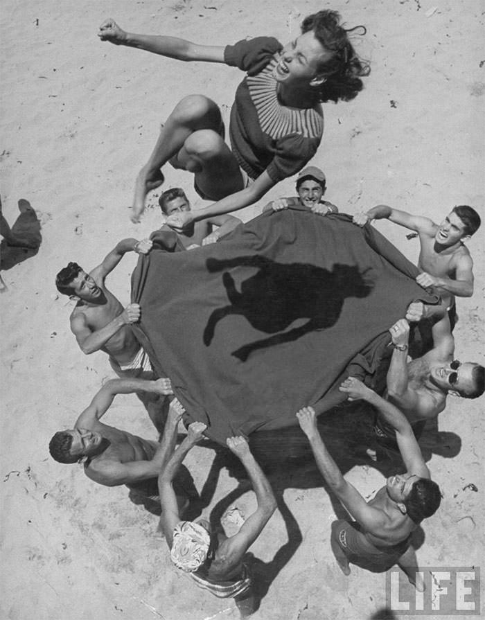 Teenaged boys using blanket to toss their friend, norma baker, into the air on the beach, 1948