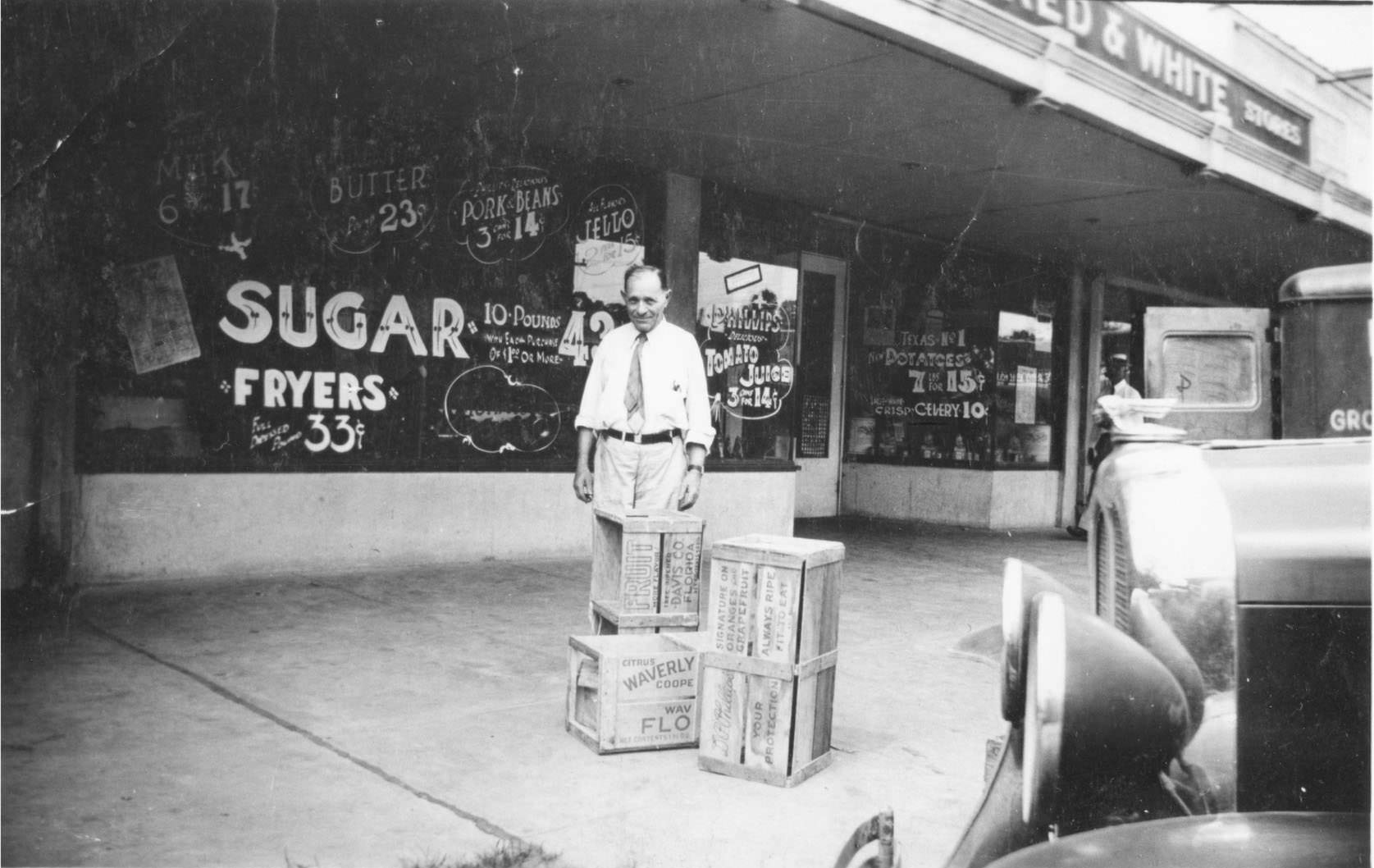 Man in front of a store with wooden crates and car in foreground.