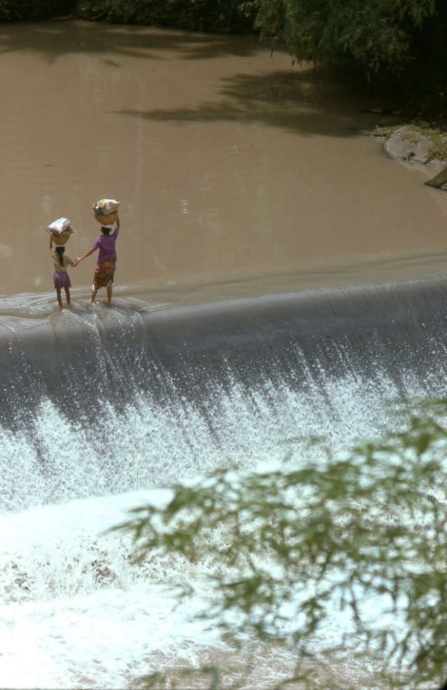 A mother leads her young daughter by the hand across a levy that spans a stream inland from Tanah Lot, 1970s