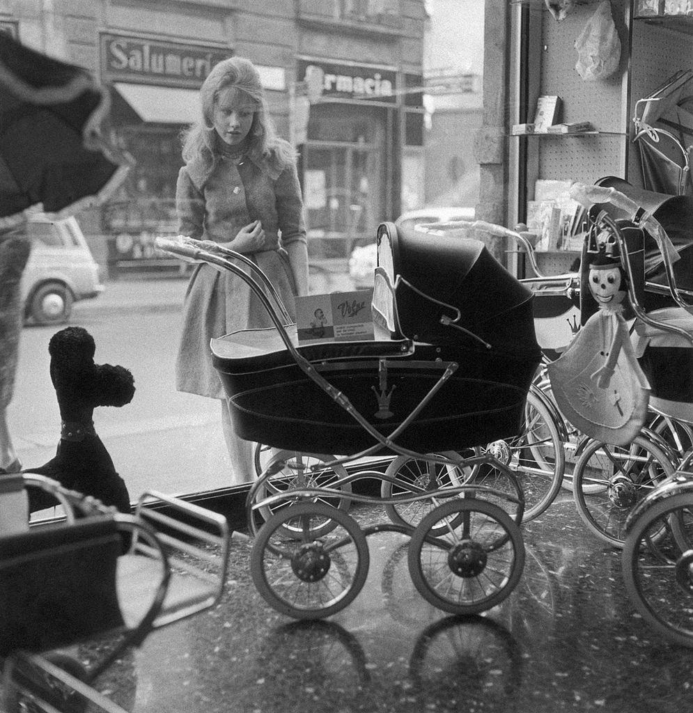 A girl looking at a pram displayed in a shop window, Italy, 1960s.