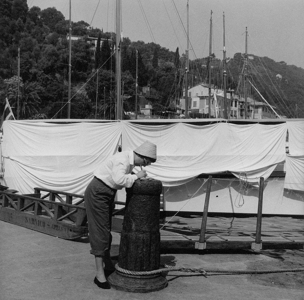 A tourist signs a postcard on a stone bollard at the dock, 1960s.