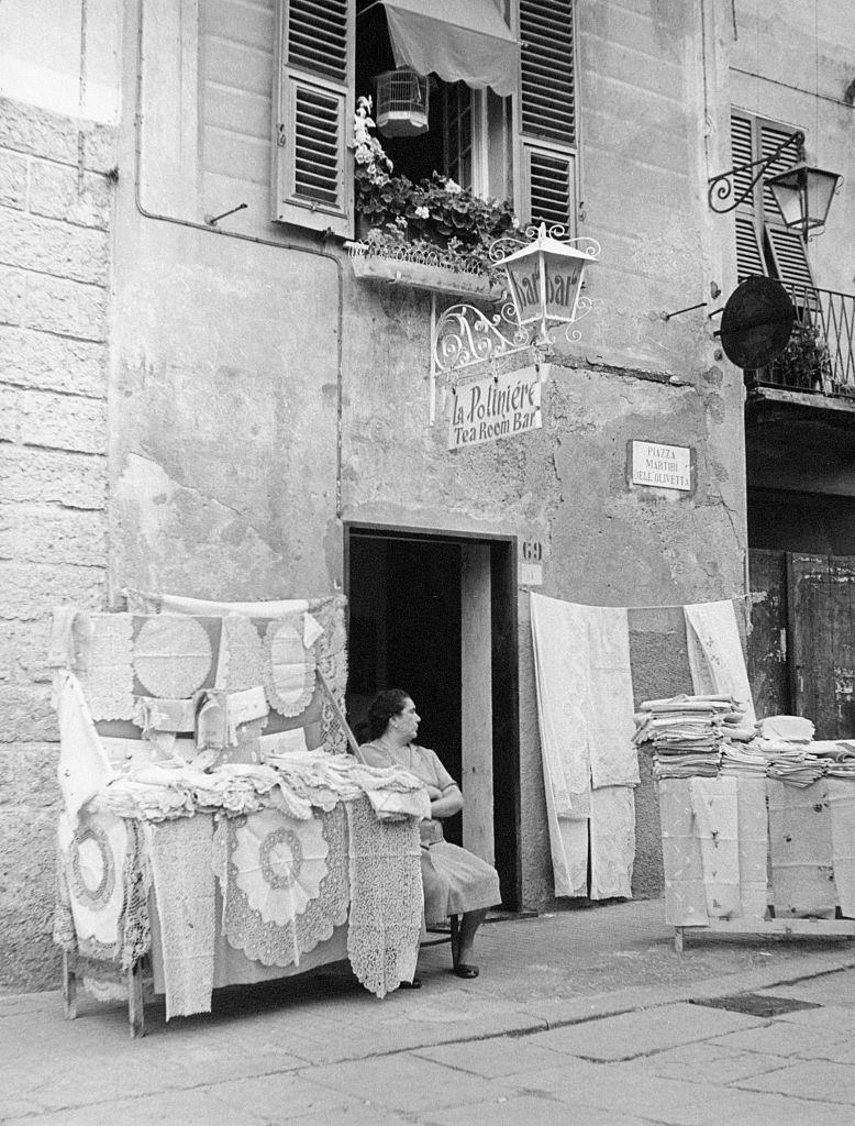 A woman sits at the entrance of the bar La PoliniÃ¨re, next to a stand of laces, 1960s.