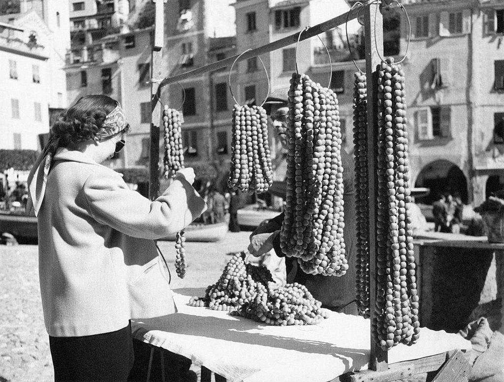 A tourist takes a look on hazelnuts necklaces exposed in a stand, Portofino, 1960s.