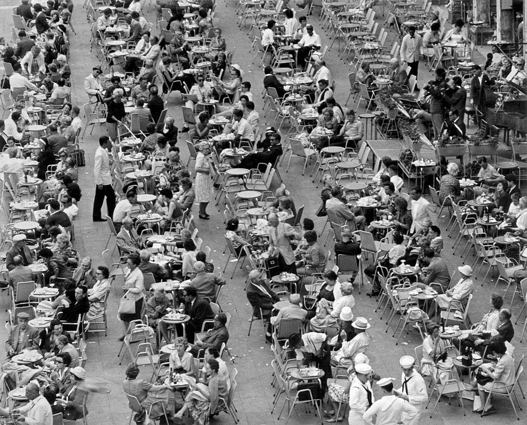 A crowd of people sitting outdoor at the tables of a café. Italy, 1960s