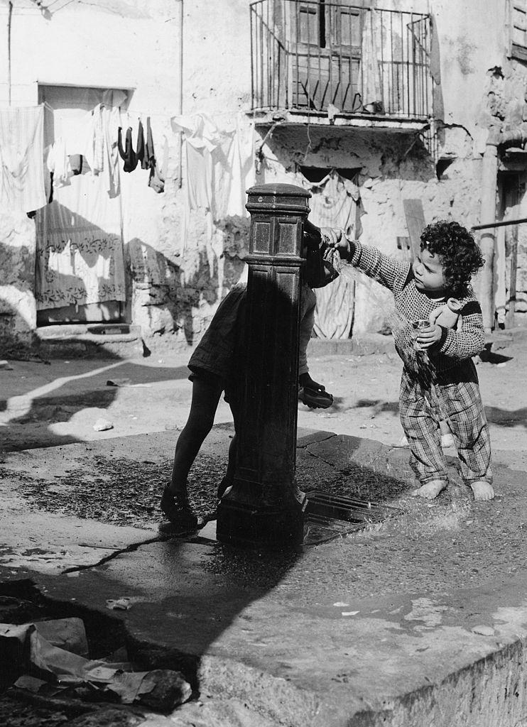 A young child at a water fountain in a slum courtyard in Italy, 1960s.