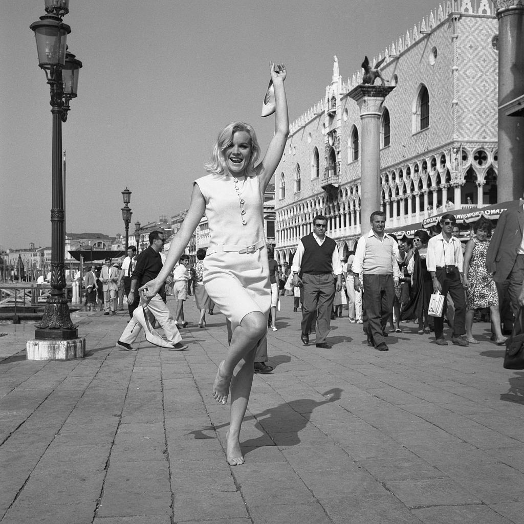 American actress Carroll Baker, wearing a white dress in Riva degli Schiavoni while standing on her tiptoe, barefoot and holding her shoes, Venice, 1960s.