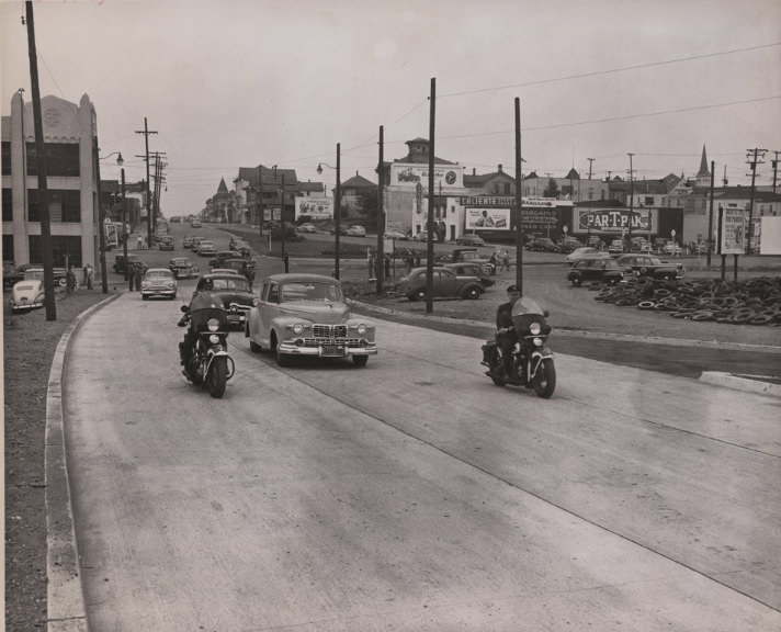 Two policeman on motorcycles escort cars eastbound on East 12th Street, 1940s