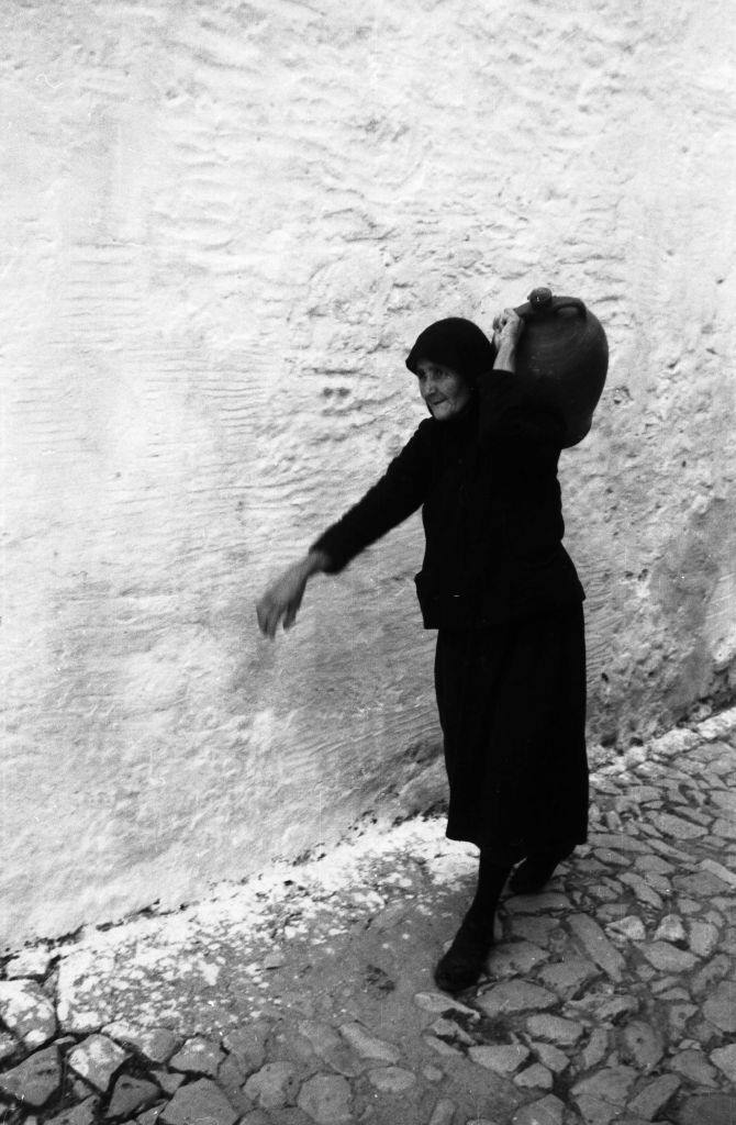An elderly woman carries a jug down an alley in Rhodes, Greece 1950s.