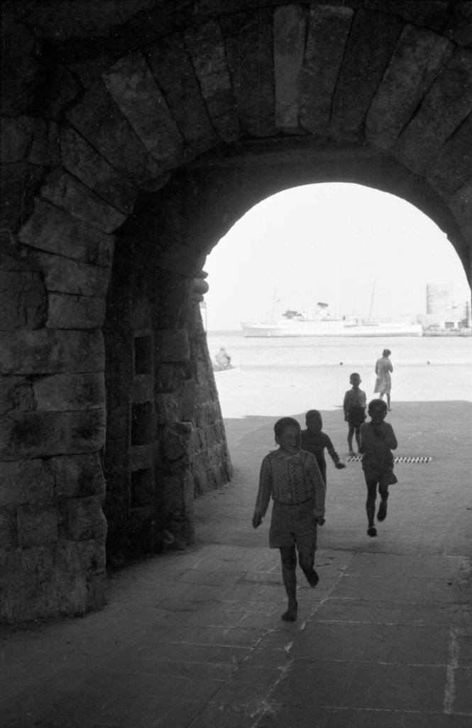 Children run from the harbor through the city walls of Rhodes to play, Greece, 1950s
