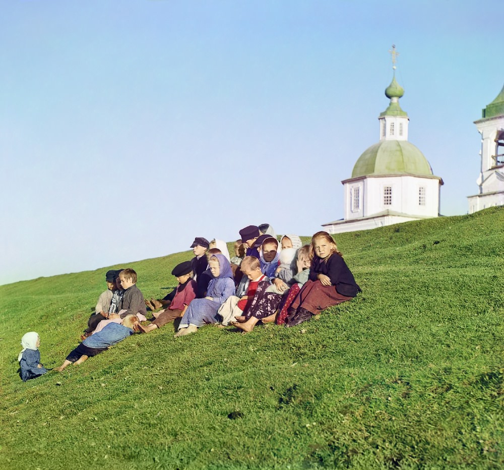 Russian children sit on the side of a hill near a church and bell tower near White Lake, in Russia, 1909.