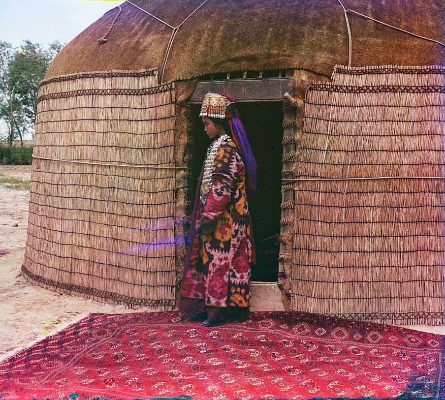 A woman standing on a carpet at the entrance to a yurt, dressed in traditional clothing and jewelry, 1910s