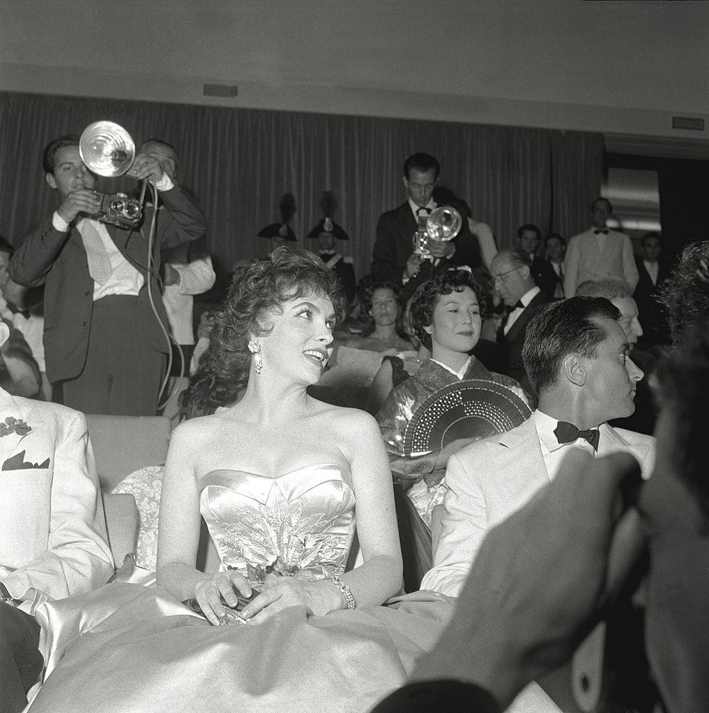 Gina Lollobrigida and her husband Milko Skofic are seated in the stalls waiting for the start of XVII Venice International Film Festival, around them photographers and members of the audience. Venice, 1956.