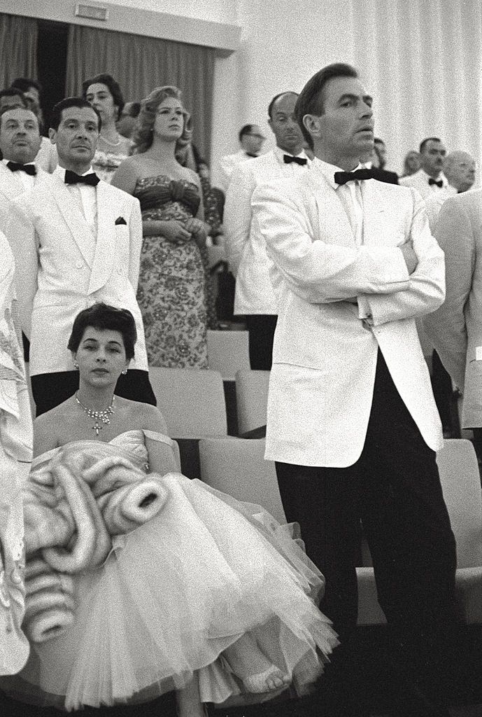 James Mason waits standing with his arms crossed, his wife Pamela Kellino is seated next to him with an annoyed look.