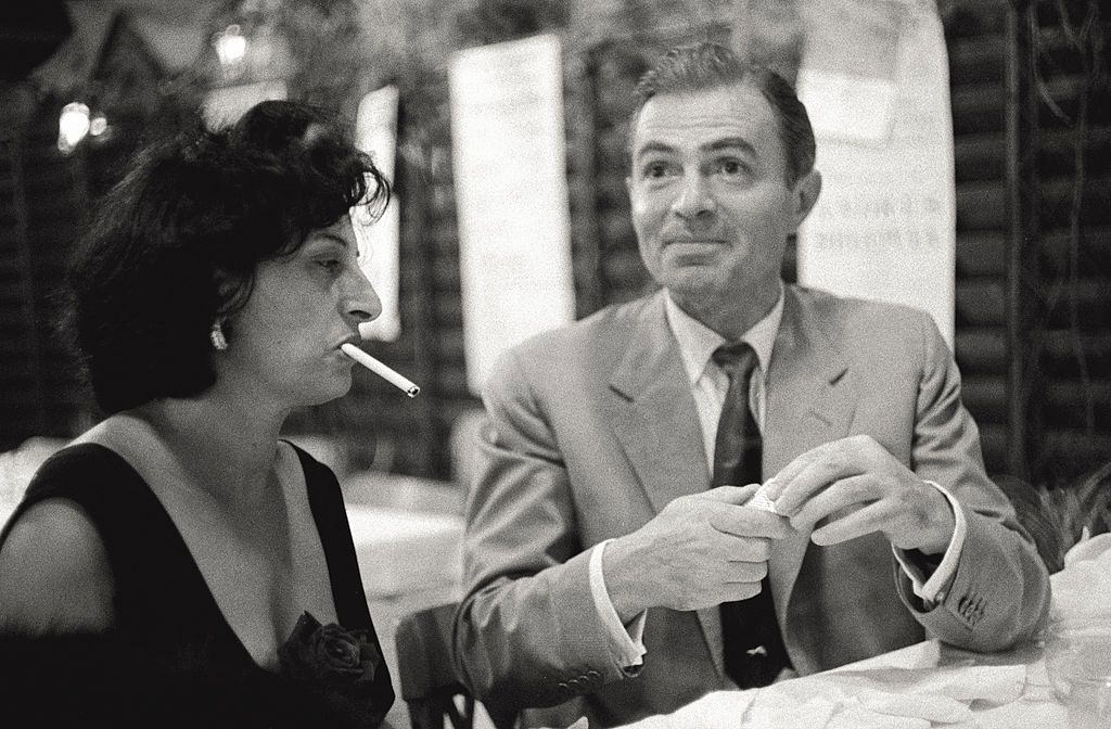The actors Anna Magnani and James Mason make conversation together, sitting at a table, during the 17th Venice Intenational Film Festival; she is waiting for him to light a cigarette.