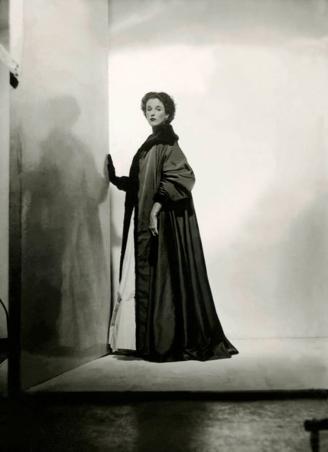Babe Paley standing with hand against a wall, and wearing a dark gray floor-length faille robe, 1946